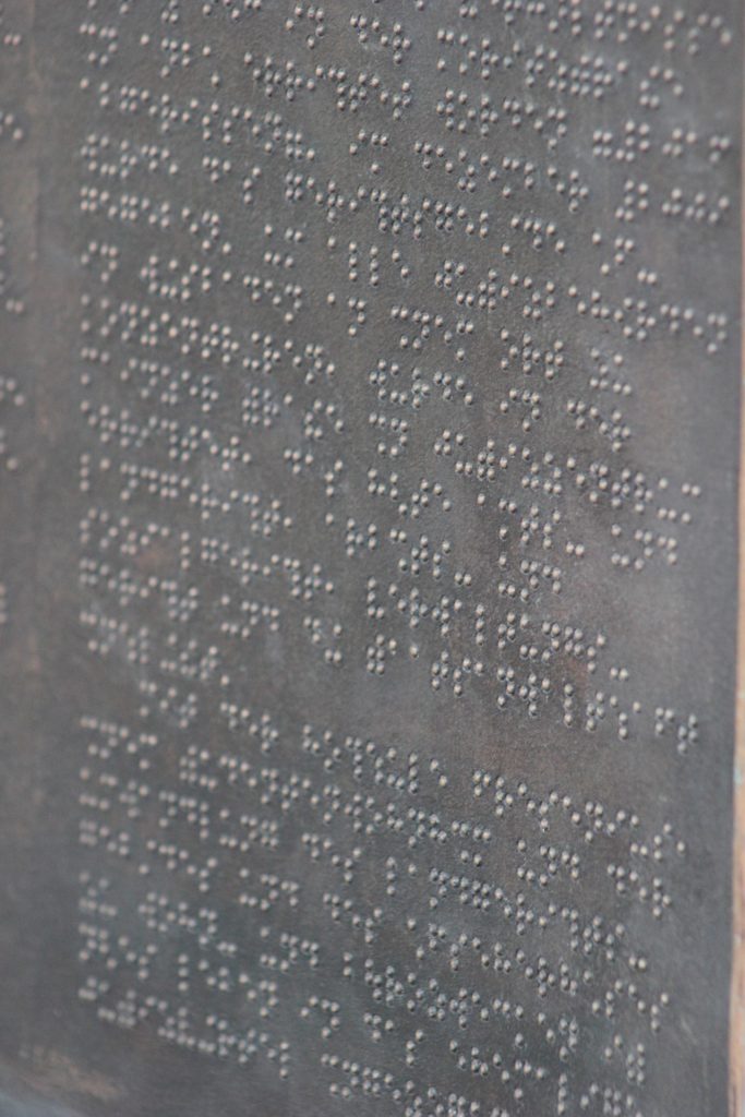 A imagem exibe uma placa cinza com texto escrito em Braille.