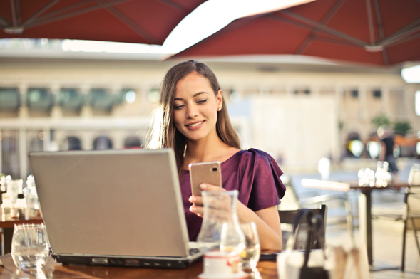 Aplicativo personalizado: Imagem de uma mulher com uma blusa roxa sentada em um restaurante olhando para seu celular. Sobre a mesa, um  computador portátil. 